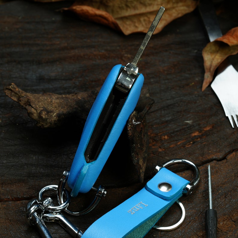 Puede incluir: Un llavero de cuero azul con un porta llaves plateado y una correa de cuero azul. El llavero tiene un anillo de llave plateado y un gancho de llave plateado.