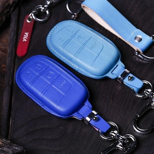 May include: A close-up of several car key fobs in various shades of blue. The key fobs are made of leather and have silver key rings. One key fob is a deep blue, and another is a lighter shade of blue. A red tag with the word "VILA" is also visible.