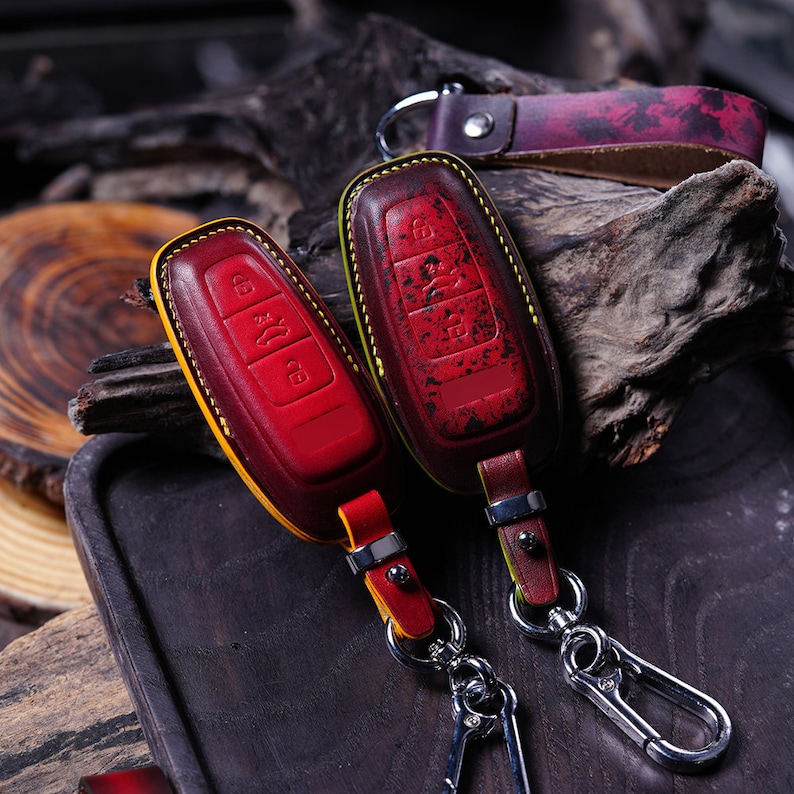 May include: Two red and yellow leather key fob covers with silver key rings. The key fobs have a distressed finish and feature button designs. A matching leather strap with a key ring is also visible.