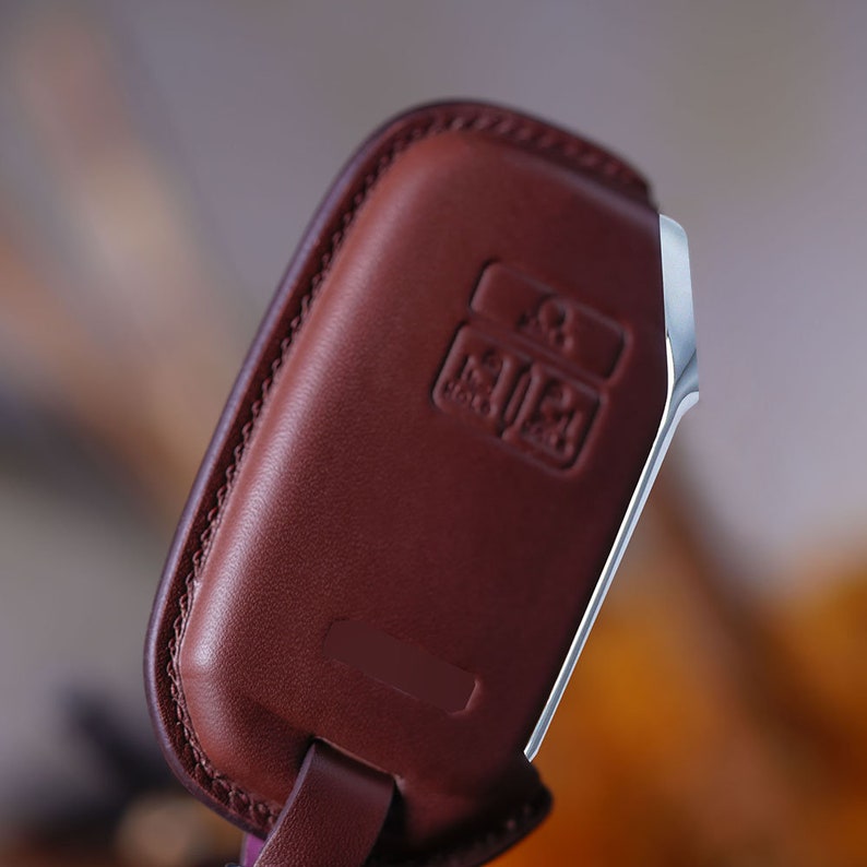 May include: A brown leather key fob with a silver metal trim. The fob has a small rectangular button with a logo on it.