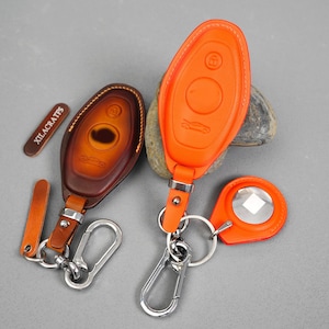 May include: Two leather key fob covers in brown and orange, with silver key rings and clasps. The brown fob has a car silhouette and a keyhole design. The orange fob has a car silhouette and a lock symbol. A small leather tag reads "XILACRATES".