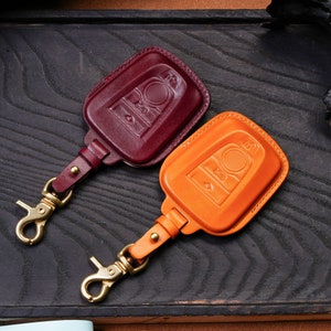 May include: Two leather keychains with a key fob design. One keychain is burgundy and the other is orange. Both keychains have a gold metal clasp.