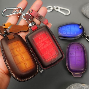 Könnte beinhalten: Leder-Schlüsselanhänger in Braun, Rot, Blau und Lila. Die Schlüsselanhänger haben einen Metallverschluss und einen Lederriemen. Die Schlüsselanhänger dienen zum Schutz von Autoschlüsseln. Die Schlüsselanhänger haben eine Vielzahl von Knopfanordnungen.