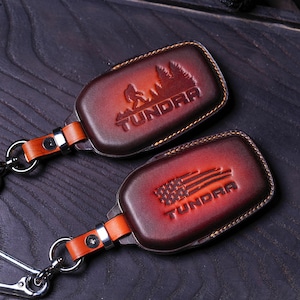 May include: Two brown leather keychains with the word "Tundra" embossed on them. One keychain features a silhouette of a person and trees, while the other features a stylized American flag.
