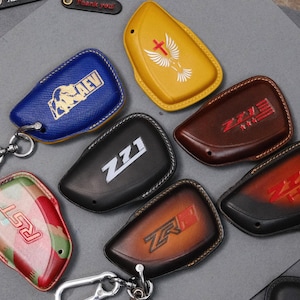 May include: A selection of leather key fob covers in various colours, including blue, yellow, brown, black, and camouflage. Each cover features unique designs and logos, with keychains attached. The covers are designed to protect car keys.