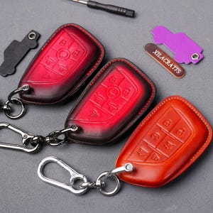 Puede incluir: Tres llaveros con fundas de cuero para llaves en rojo, naranja y marrón. Cada funda tiene un diseño de botones diferente. Los llaveros tienen anillos y ganchos de metal plateado.