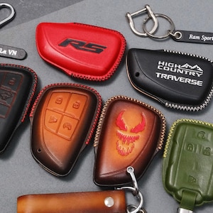 May include: A collection of leather key fob covers in various colors, including red, black, brown, and green. Some covers feature embossed designs and text, such as "RS", "High Country Traverse", and a stylized face. Keychains and key tags are also included.