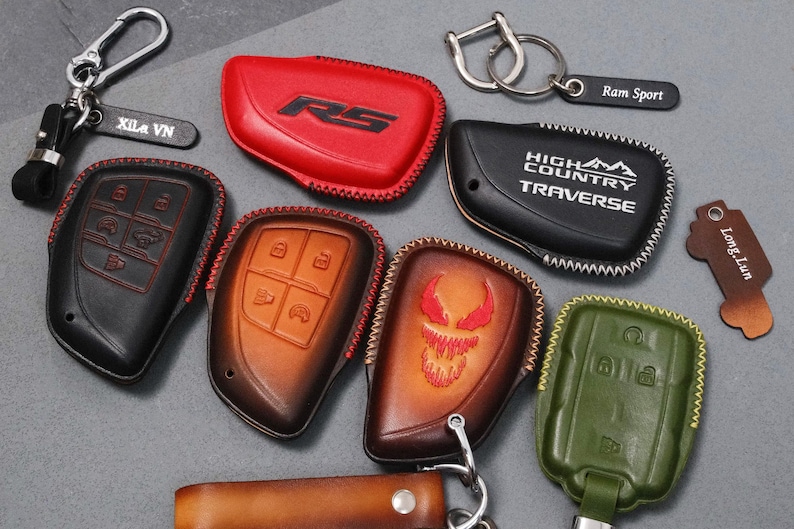 May include: A collection of leather key fob covers in various colors, including red, black, brown, and green. Some covers feature embossed designs and text, such as "RS", "High Country Traverse", and a stylized face. Keychains and key tags are also included.