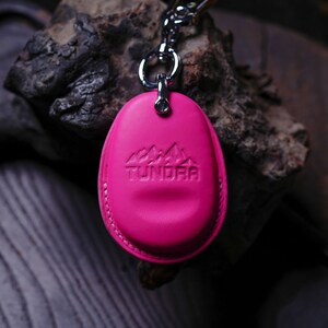 May include: A pink leather keychain with a silver ring and a silver chain. The keychain has the word "TUNDRA" embossed on it, along with a mountain range design.