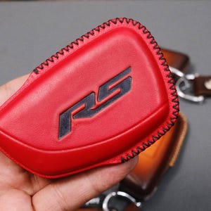 May include: A red leather key fob cover with black stitching and the letters "RS" in black. The fob is held in a hand, with other leather accessories visible in the background.
