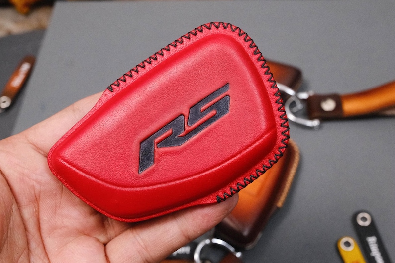 May include: A red leather key fob cover with black stitching and the letters "RS" in black. The fob is held in a hand, with other leather accessories visible in the background.