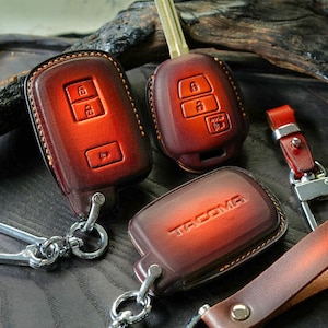 May include: Three leather keychains with a red-brown gradient. The keychains feature a key fob with buttons, a key fob with the word "TACOMA" embossed, and a key fob with a silver ring.