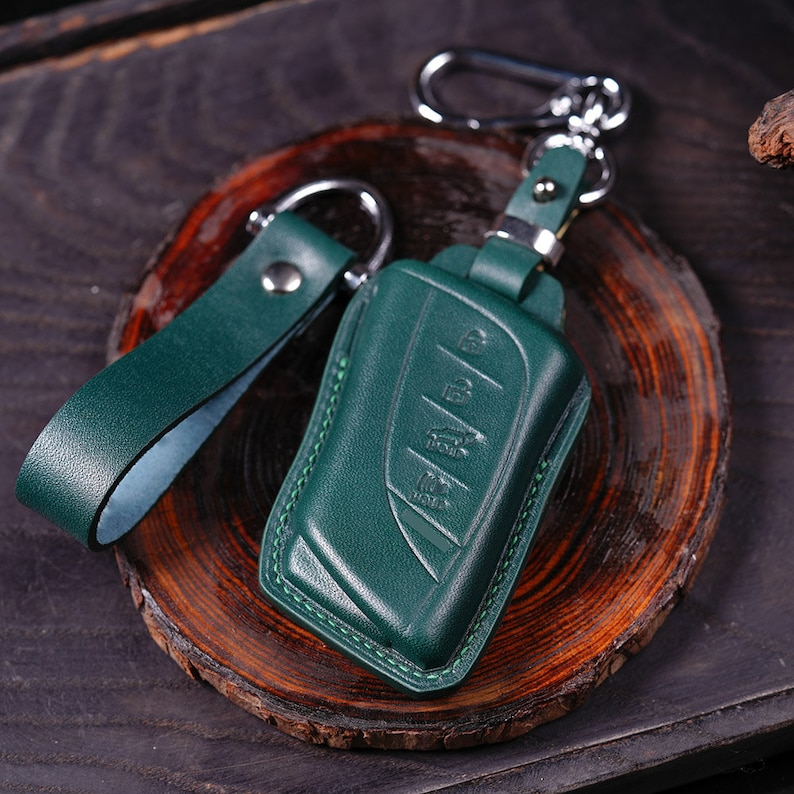 May include: A dark green leather key fob with a silver metal hook and loop. The fob has a stitched edge and a smooth surface.