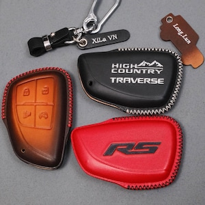 May include: A collection of leather key fob covers in various colors. Includes a brown and black cover with button icons, a black cover with "HIGH COUNTRY TRAVERSE" text, and a red cover with "RS" text. Also includes keychains.