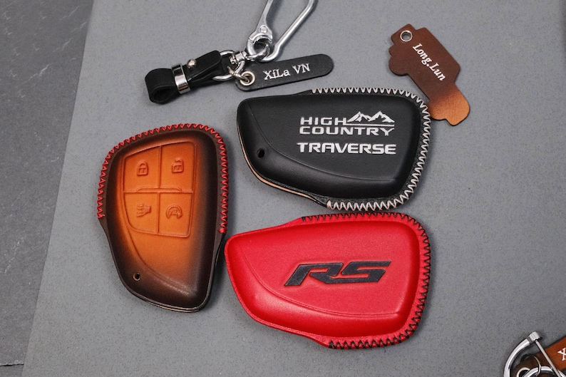 May include: A collection of leather key fob covers in various colors. Includes a brown and black cover with button icons, a black cover with "HIGH COUNTRY TRAVERSE" text, and a red cover with "RS" text. Also includes keychains.