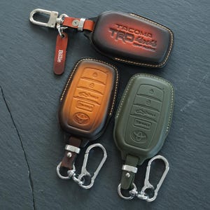 May include: Three leather key fob covers for Toyota vehicles. One is dark brown with 'TACOMA TRD 4x4 OFF ROAD' embossed, one is tan with a Toyota logo, and one is olive green with a Toyota logo. Each includes a silver metal clip and a leather tag.