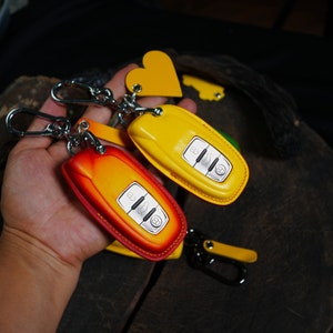 Könnte beinhalten: Zwei Schlüsselhüllen aus Leder, eine gelbe und eine orange, mit einem silbernen Schlüsselanhänger. Die Schlüsselhüllen sind an einer silbernen Kette mit einem herzförmigen Anhänger befestigt.