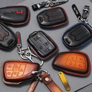 May include: A collection of leather key fob covers in various colors, including black and brown. Some have red or blue stitching. Keychains and key rings are attached to the covers. The key fobs have embossed button designs.