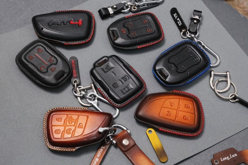 May include: A collection of leather key fob covers in various colors, including black and brown. Some have red or blue stitching. Keychains and key rings are attached to the covers. The key fobs have embossed button designs.
