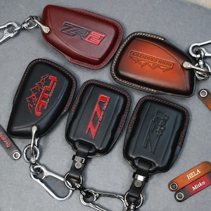 May include: A collection of leather key fobs in various colors, including black and brown. The fobs feature embossed designs and text, such as "ZR2", "AT4", and "High Country". Some fobs have silver key rings and clasps.