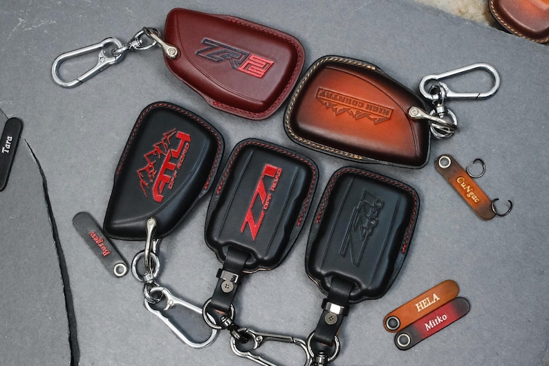 May include: A collection of leather key fobs in various colors, including black and brown. The fobs feature embossed designs and text, such as "ZR2", "AT4", and "High Country". Some fobs have silver key rings and clasps.