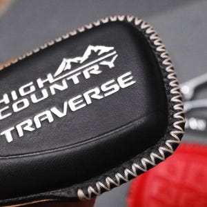 May include: Black leather key fob cover with white stitching and the words "HIGH COUNTRY TRAVERSE" in white. The cover has a mountain graphic above the text. A silver key ring and red object are visible in the background.