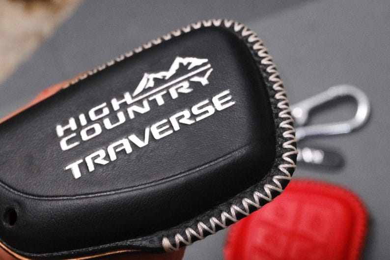 May include: Black leather key fob cover with white stitching and the words "HIGH COUNTRY TRAVERSE" in white. The cover has a mountain graphic above the text. A silver key ring and red object are visible in the background.