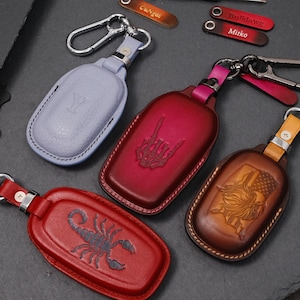 May include: A collection of leather keychains in various colors, including light blue, red, and brown. Each keychain features a unique design, such as a scorpion, a hand gesture, and a Texas-themed graphic. They have metal clasps.