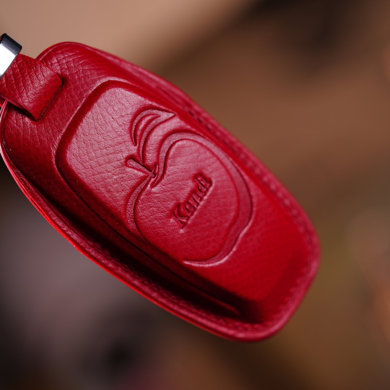 May include: A red leather key fob with a debossed apple design and the name "Kandi". The fob has a rectangular shape with rounded edges and a silver-colored metal loop for attaching keys.