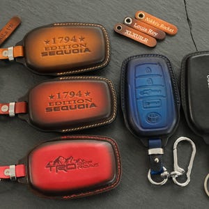 May include: A collection of leather key fob covers in various colors including tan, brown, red, blue, and black. Some covers feature embossed text such as 1794 Edition Sequoia or TRD Off Road. Small leather tags with names are included with key rings.