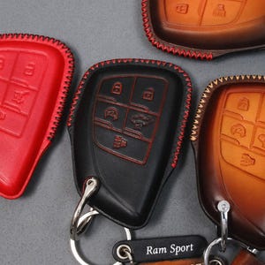 May include: Leather key fob covers in red, black, and brown. Each cover has a stitched edge and key button icons. The black cover has red stitching. The brown cover has a leather strap and a metal key ring. The text "Ram Sport" is visible.