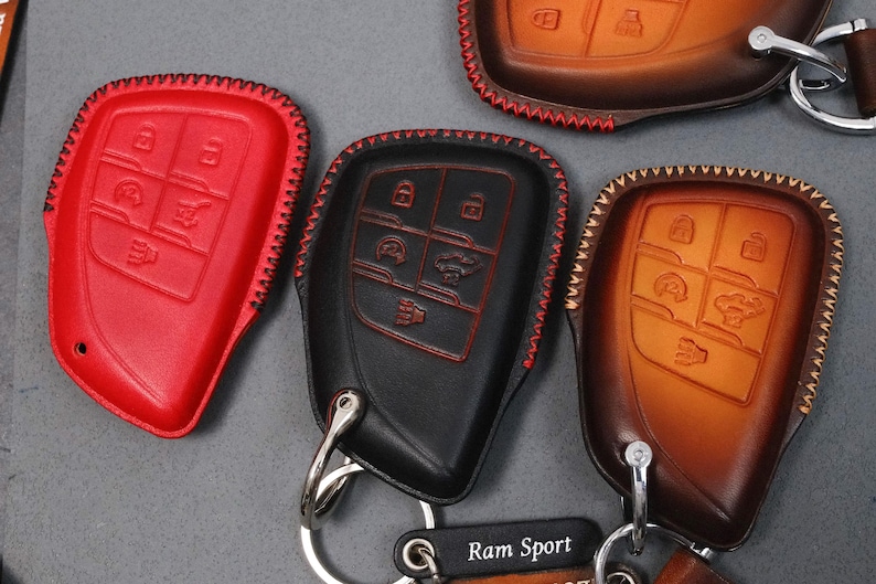 May include: Leather key fob covers in red, black, and brown. Each cover has a stitched edge and key button icons. The black cover has red stitching. The brown cover has a leather strap and a metal key ring. The text "Ram Sport" is visible.