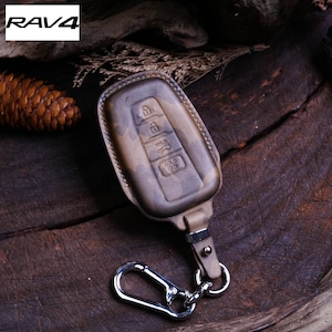 May include: A camouflage patterned leather key fob with a silver key ring and a car key with buttons labeled "HOLD" and "HOLD 1/3". The key fob is on a wooden surface.