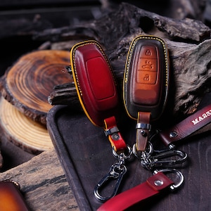 May include: Two leather key fob covers in red and brown, with yellow stitching. The key fobs have silver key rings and leather straps. The background is a dark wooden surface.