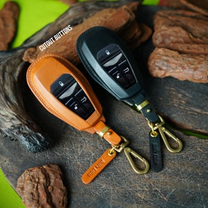 May include: Two leather key fobs with cutout buttons and a gold metal carabiner. The key fobs are brown and black leather. The text "CUT OUT BUTTONS" is visible in the top left corner of the image.