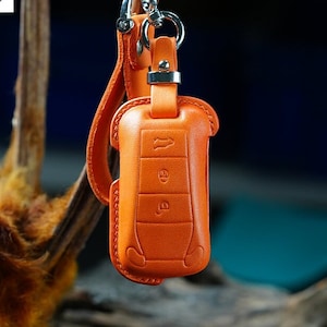 Puede incluir: Llavero de cuero naranja con un anillo y una correa plateados. El llavero tiene cuatro botones con símbolos en relieve.
