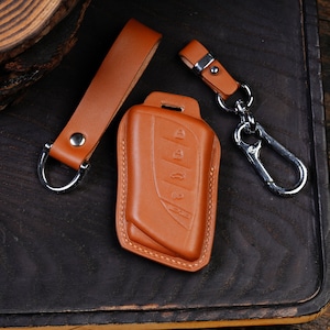 May include: A tan leather key fob cover, key strap, and keychain set. The key fob cover has embossed buttons and the brand name "LEXUS". The set includes a silver-tone carabiner clip and a matching leather strap with a metal ring.