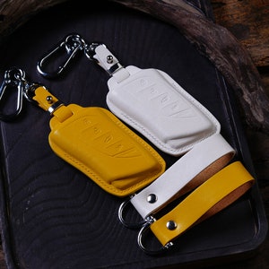 Peut inclure: Deux porte-clés en cuir avec des anneaux métalliques et des housses de clés de voiture. Un porte-clés est jaune et l'autre est blanc.