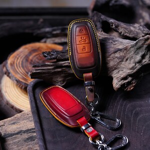 May include: Two leather key fob covers in shades of brown and red, with yellow stitching and silver key rings. The key fobs have a leather strap and metal clasp. The background features wood and a dark tray.