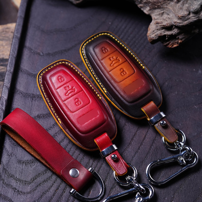 May include: Two leather car key fobs in red and brown, with yellow stitching and silver keychains. The red fob has a matching leather strap. The fobs have button icons for locking, unlocking, and other car functions.