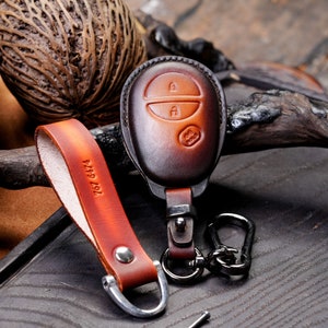 Puede incluir: Un llavero de cuero marrón con un anillo de llave plateado y un lazo para sujetarlo a un cinturón. El llavero tiene un diseño de ojo de cerradura y el texto "767 6474" está grabado en el lazo.