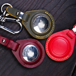 Puede incluir: Tres llaveros de cuero con anillos metálicos y un recorte circular en el centro. Los llaveros son verde, rojo y burdeos. El texto "Ultra Wideband AirTag - Designed by Apple in California" está grabado en el llavero verde.