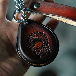 May include: A teardrop-shaped, dark brown leather keychain with a skull wearing a Native American headdress embossed on the front. The keychain has a silver metal clasp and a matching leather strap. The edges are stitched with orange thread.