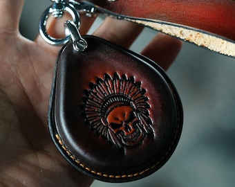 Puede incluir: Llavero de cuero marrón oscuro en forma de lágrima, con una calavera en relieve con un tocado nativo americano. Tiene un cierre de metal plateado y una correa de cuero a juego. Los bordes están cosidos con hilo naranja.