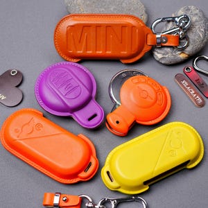 May include: Four different key fob covers in various colors, including orange, yellow, purple, and brown. The covers are made of leather and have a variety of designs. The word "MINI" is embossed on the brown cover.