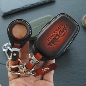 May include: A brown and black leather key fob cover for a Toyota Tacoma TRD 4x4 Off Road vehicle, featuring a circular cutout for a key fob, a metal clip, and a small rectangular leather tag with the name Billie embossed in white lettering.