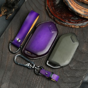 May include: A collection of leather key fobs and key cases in shades of purple and olive green. The key fobs have silver hardware and a matching purple leather strap. The key cases are designed to protect car keys.