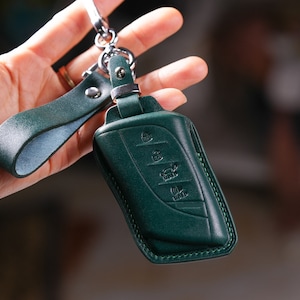 May include: A dark green leather key fob with a silver chain and a loop for attaching to a keychain. The fob has a stitched edge and embossed buttons.