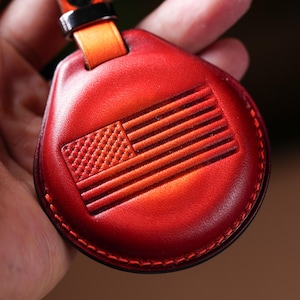 May include: Red leather keychain with an embossed American flag design. The keychain has a stitched border and an orange strap with a black metal loop. The leather has a gradient effect, transitioning from a darker red to a lighter shade.