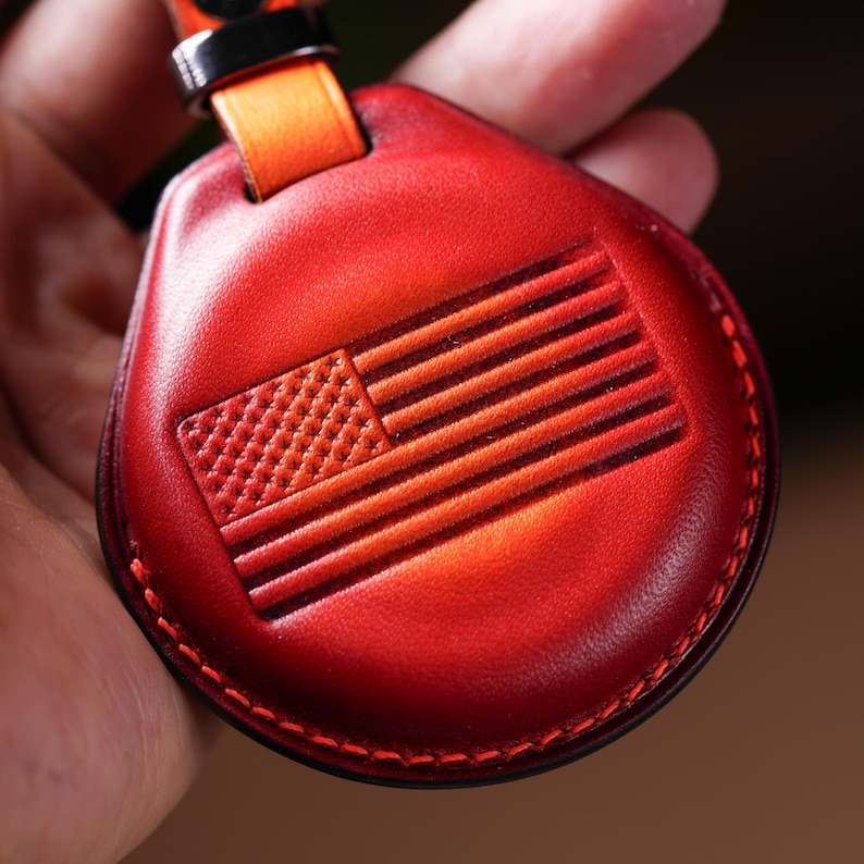 May include: Red leather keychain with an embossed American flag design. The keychain has a stitched border and an orange strap with a black metal loop. The leather has a gradient effect, transitioning from a darker red to a lighter shade.